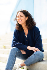 woman with curly brown hair sitting on outdoor steps, looking ahead with a smile
