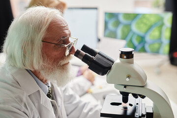 Senior Caucasian man with grey hair and beard using microscope in scientific laboratory, focusing...