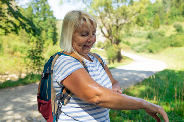 Smiling senior woman with a backpack applying cream or insect repellent on her arm during a summer...
