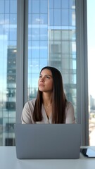 Contemplative young professional woman works at a laptop by the office window