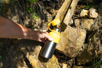 Close-up of a hand collecting fresh water from a mountain spring into a reusable bottle. Captures...