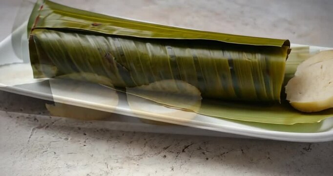 A long, light-colored food item wrapped in large, green leaves, sliced and on a white rectangular plate