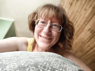 Middle-aged woman with disheveled hair smiles for a selfie in cozy bedroom after waking up