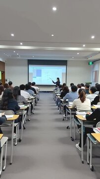 A teacher professor is pointing at a monitor gives a lecture lesson to students sitting in a spacious classroom, vertical video