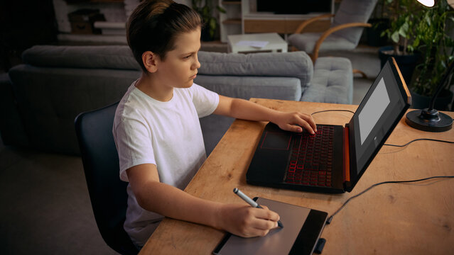 Boy uses a pen and tablet for digital drawing or design while interacting with a laptop. Concept of digital creativity, graphic design training and graphic design training