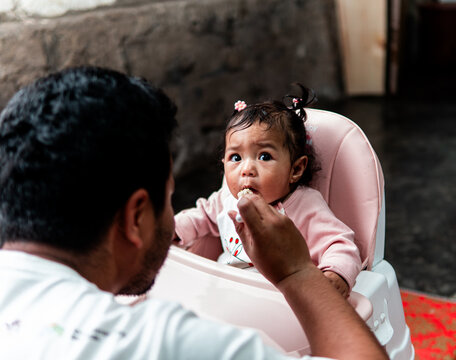 A cute baby girl sitting in a high chair looking curious while her father feeds her with a spoon. A heartwarming family moment of love and care at home.