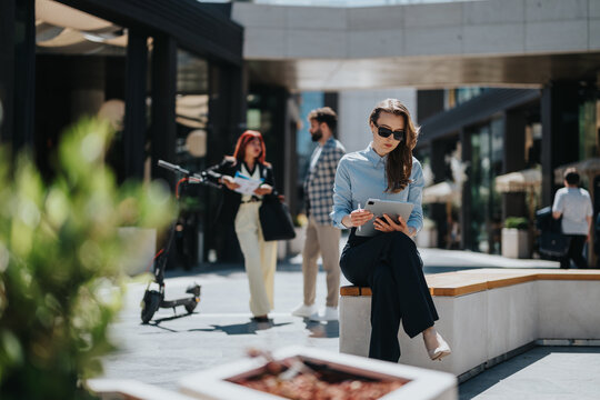 A confident businesswoman using a tablet in an urban environment surrounded by colleagues. The scene highlights modern professionalism, technology integration, and collaboration in an open workplace.
