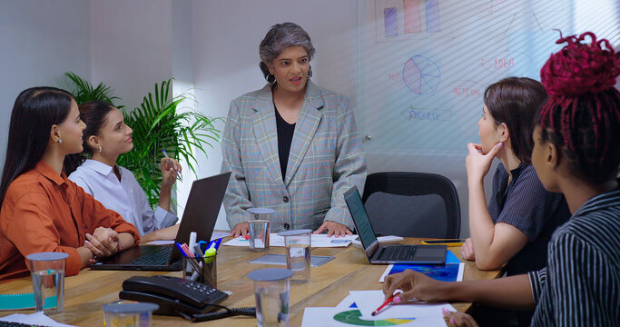 Indian old senior boss woman lady talk speak show white board chart learn study graph data growth work place. young adult female girl staff group team share stock trade new day idea plan job office
