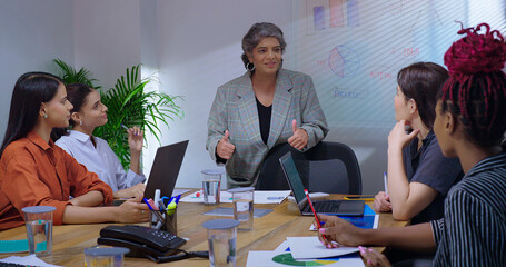 Indian old senior boss woman lady talk speak show thumb up white board chart learn study graph data work place. young adult girl staff group team share stock trade new day idea plan job office