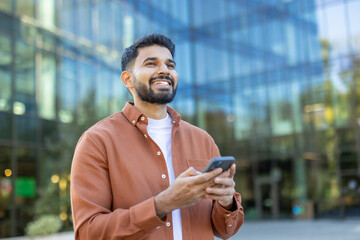 A smiling man holds a smartphone in front of a modern glass building, suggesting a business or...