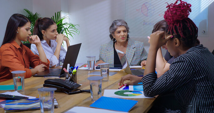 Indian young adult woman staff sitting around desk table with old aged boss ceo lady talk share bad news at job place upset female leader speak about work loss to girl group team office board room - Powered by Adobe