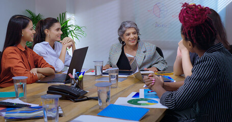 Indian young adult woman group sitting around table desk chair with boss CEO lady talk gossip enjoy work day indoor office female girl staff team group speak share good news spend break time job place
