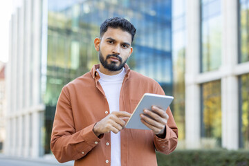 A man with a beard uses a tablet in front of a modern building, looking directly at the camera.