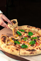 Woman’s Hand Lifting a Slice of Wood‑Fired Neapolitan Pizza on Rustic Table. Fresh, savory moment perfect for restaurant ads, menu visuals and food blogs.