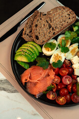 Hearty Smoked Salmon Breakfast Bowl with Avocado, Egg & Feta. Multigrain bread, cherry tomatoes, cucumber and olives plated for a nutritious morning meal.