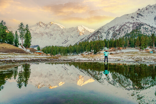 A traveler stands joyfully at Fairy Meadows, Pakistan, with Nanga Parbat rising behind. Snow, pine trees, and a perfect lake reflection create a serene adventure scene.