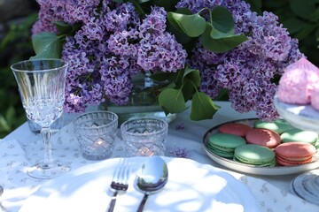 Beautiful lilac flowers, sweets, plates, cutlery, glass and burning candles on table in garden,...