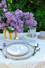 Beautiful lilac flowers, plates, cutlery, glasses and candles on table in garden