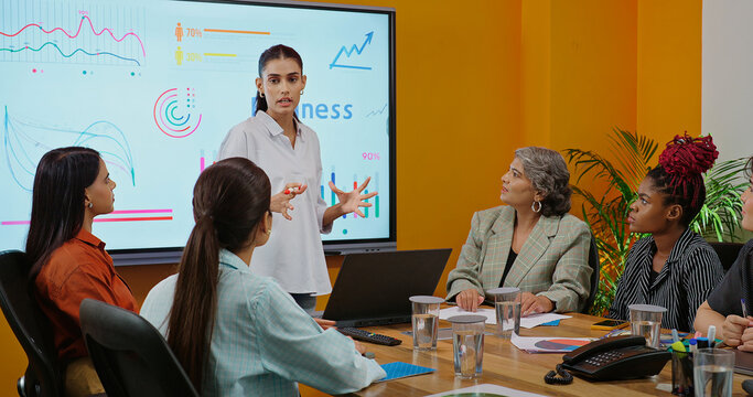 Indian young female old boss lady team leader show chart white board learn study graph data growth serious work place Asian adult girl staff group talk speak share stock trade idea plan job office day