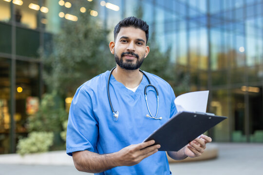 A medical professional, likely a doctor or nurse, stands outside a building, holding a clipboard and looking at the camera.