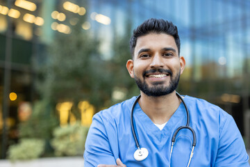 A smiling male doctor in blue scrubs stands in front of a modern building, radiating confidence and...
