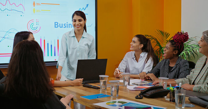 Indian young female old boss lady team leader show chart white board learn study graph data growth work place adult girl talk speak share stock trade idea plan staff group job office day