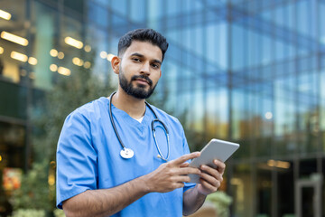 A healthcare professional in scrubs uses a tablet outdoors, in front of a modern building. He wears...