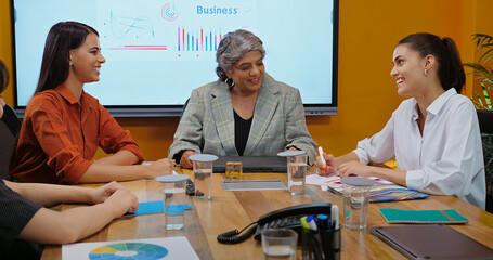 Indian smiling old boss ceo woman lady show stock data chart paper growth talk share ideas plan young adult expert girl staff group intern team speak do work day task make new deal at job place