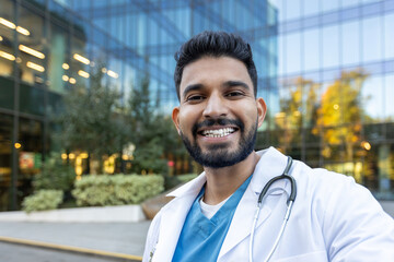 A smiling doctor stands in front of a modern building, wearing a lab coat and stethoscope. He looks...