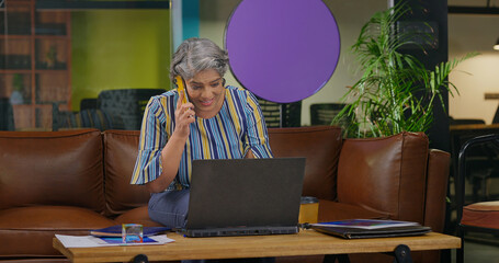 Indian older woman lady sitting on sofa couch use laptop look screen do online work check bills e-bank data app Asian happy adult aged female boss talk speak on phone call share good news at office
