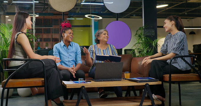 Indian old aged boss lady hold file do share good news new deal work talk speak ideas plan job place happy female girl staff group team sitting on chair sofa couch use laptop look screen at office