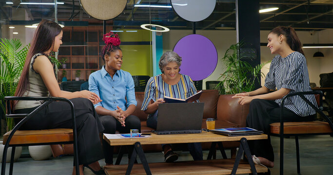 Indian old aged boss lady hold file do share good news new deal work talk speak ideas plan job place happy female girl staff group team sitting on chair sofa couch use laptop look screen at office