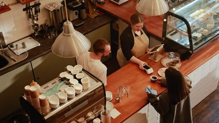 A female customer is talking to a male employee and a female owner standing across from her at the bar, she takes a cup and pays with the terminal