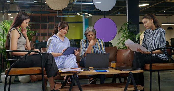 Indian serious busy woman lady boss sitting sofa couch chair desk table use laptop do work online email young adult female girl team expert staff hold paper read check report day time at job place	