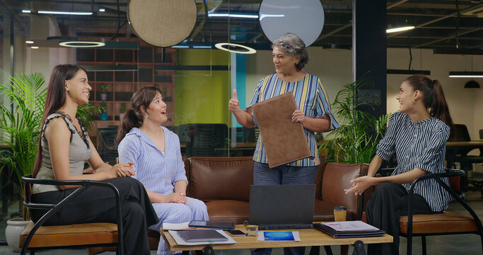 Indian old aged boss lady hold file do share good news new deal work talk speak ideas plan job place happy female girl staff group team sitting on chair sofa couch clap hand show thumbs up at office