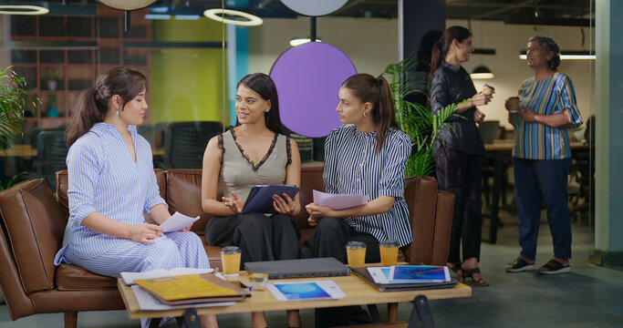 Indian happy busy woman leader young adult staff sitting sofa use tablet look screen check email talk work day task with team female girl expert hold paper listen share new ideas plan job office place