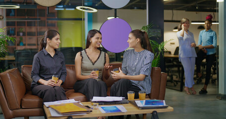 Indian asian smiling young adult gen z diverse girl woman sitting on sofa drink coffee indoor job office happy female intern staff talk funny gossip spend break time enjoy fun joy day at work place