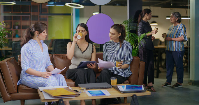 Indian young adult gen z woman lady group sitting on sofa hold cup drink coffee indoor job office female girl intern staff talk check paper work spend break time enjoy have fun joy day at job place
