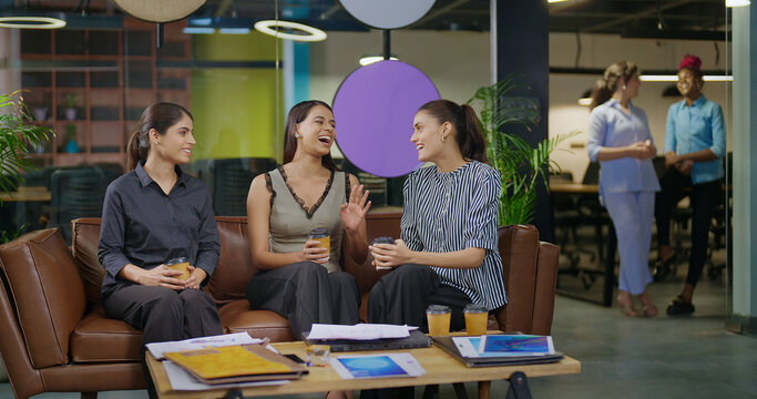Indian asian smiling young adult gen z diverse girl woman sitting on sofa drink coffee indoor job office happy female intern staff talk funny gossip spend break time enjoy fun joy day at work place