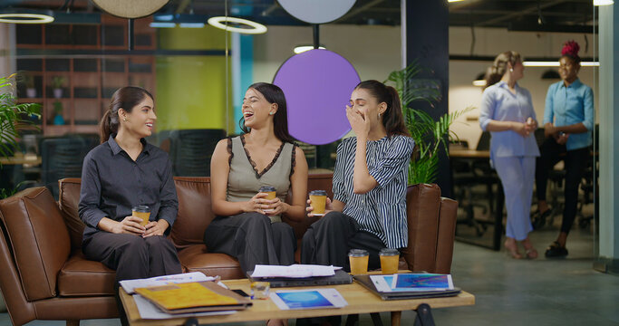 Indian asian smiling young adult gen z diverse girl woman sitting on sofa drink coffee indoor job office happy female intern staff talk funny gossip spend break time enjoy fun joy day at work place - Powered by Adobe