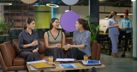 Indian asian smiling young adult gen z diverse girl woman sitting on sofa drink coffee indoor job office happy female intern staff talk funny gossip spend break time enjoy fun joy day at work place