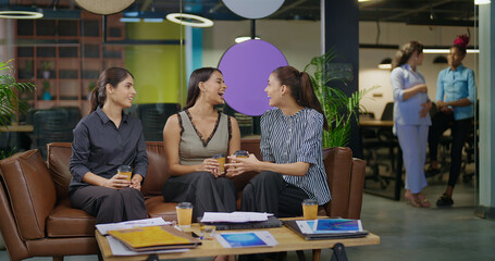 Indian asian smiling young adult gen z diverse girl woman sitting on sofa drink coffee indoor job office happy female intern staff talk funny gossip spend break time enjoy fun joy day at work place