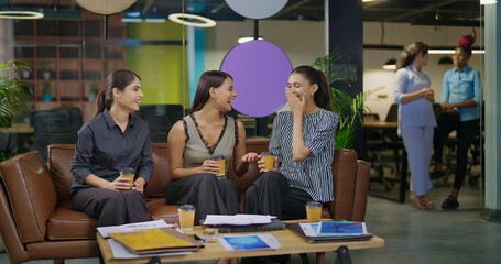 Indian young adult gen z woman lady group sitting on sofa hold cup drink coffee indoor job office happy female girl intern staff talk funny gossip spend break time enjoy fun joy day at job work place
