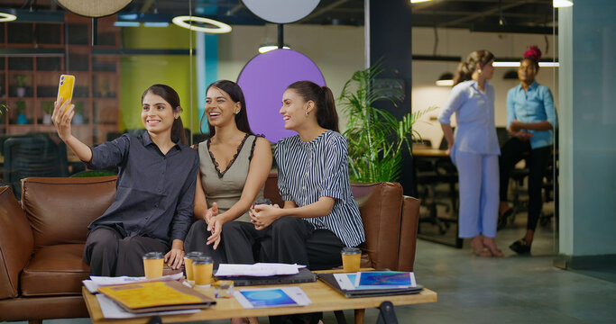 Indian happy young adult gen z lady sitting cozy sofa hold mobile look cam take selfie casual pose job work place teen girl use phone make video shoot enjoy office day have fun joy break time - Powered by Adobe