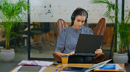 Indian young adult gen z busy woman lady sitting on chair wear headphone use laptop look screen do...