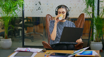 Indian young adult gen z busy woman lady sitting on chair wear headphone use laptop look screen do...