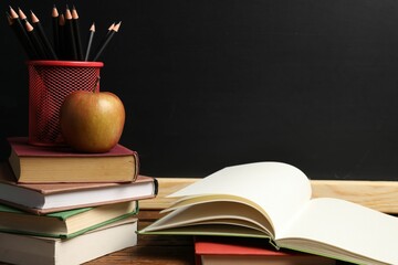 Different books, pencils and apple on wooden table near blackboard, closeup