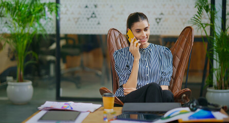 Indian young adult gen z woman lady sitting on chair hold mobile talk gossip enjoy call indoor job...