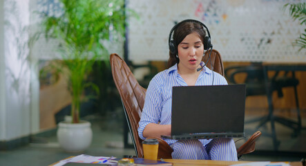 afghan young adult gen z busy woman lady sitting on chair wear headphone use laptop look screen do...