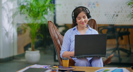 afghan young adult gen z busy woman lady sitting on chair wear headphone use laptop look screen do...